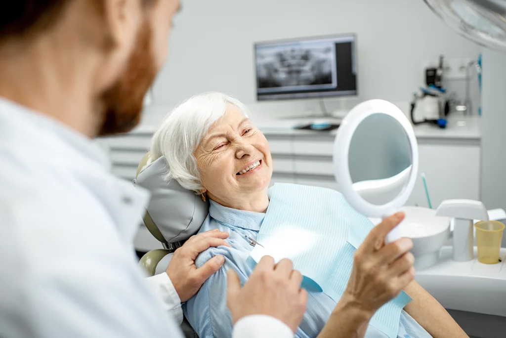 Elderly woman smiling while holding up a mirror to see her new teeth. Dentist is next to her.
