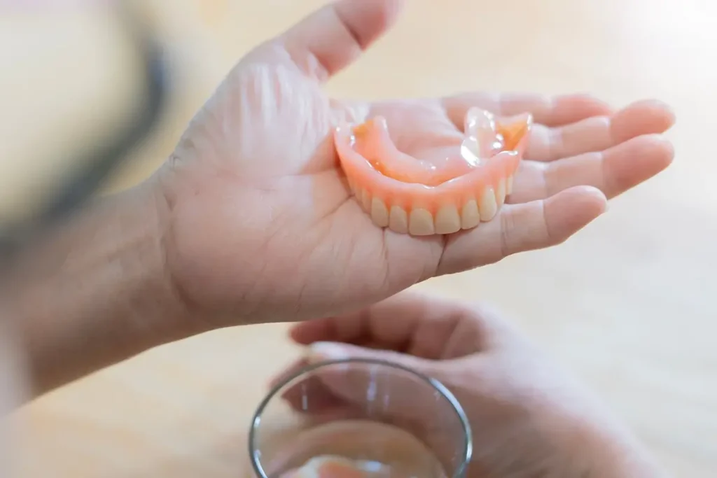 Hand holding top dentures other hand underneath on table next to a glass.